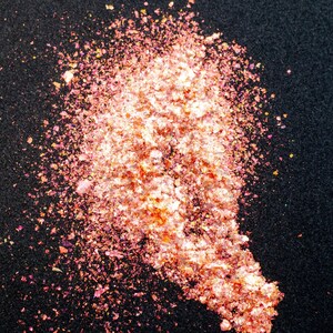 May include: A pile of iridescent, metallic flakes in shades of peach, copper, and gold. The small, irregular, reflective pieces are scattered in a dense, elongated cluster, creating a shimmering, multi-dimensional texture with a high-gloss finish.