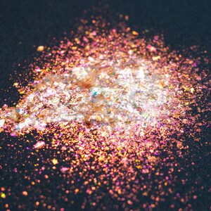 May include: A pile of iridescent, multi-colored craft flakes featuring a mix of gold, pink, and orange hues. The thin, reflective particles have a metallic finish and are scattered across a dark surface, creating a shimmering, light-catching effect.