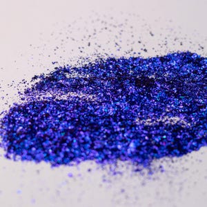 May include: A pile of loose, iridescent dark blue and violet craft flakes with a metallic finish. The small, irregular particles are scattered across the surface, creating a dense, shimmering cluster that reflects light with deep blue tones.