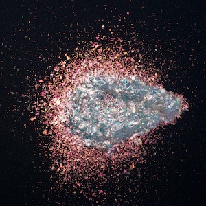 May include: A loose pile of iridescent cosmetic glitter featuring a central cluster of light blue and silver reflective flakes surrounded by a scattered halo of fine pink, magenta, and copper-toned metallic particles against a solid black background.