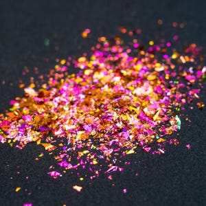 May include: A collection of small, irregular metallic flakes in vibrant shades of magenta, hot pink, and gold. These reflective, thin pieces are scattered in a loose pile, creating a shimmering effect with varying light reflections across the surface.