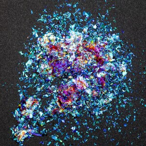 May include: A collection of iridescent, metallic flakes in shades of blue, purple, and magenta. These thin, reflective fragments are scattered in an irregular cluster, displaying a high-gloss finish that catches light to create a vibrant, multi-colored effect.
