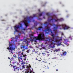 May include: A pile of loose, iridescent cosmetic pigment powder featuring deep purple, vibrant blue, and magenta flecks. The particles have a reflective, metallic finish that creates a multi-dimensional color shift across the scattered texture.