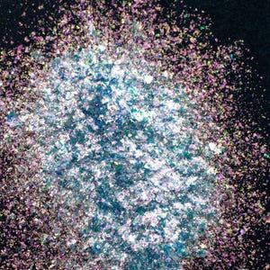 May include: A pile of iridescent, multi-colored craft flakes featuring a reflective finish. The small, thin, irregular pieces display shifting hues of blue, purple, pink, and silver, creating a shimmering, metallic effect against a dark background.
