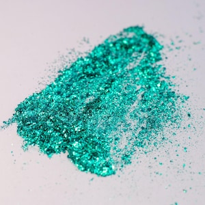 May include: A pile of loose, vibrant emerald green metallic flakes with a highly reflective, shimmering texture. The particles vary in size and shape, creating a dense, irregular cluster that catches the light to produce bright highlights.