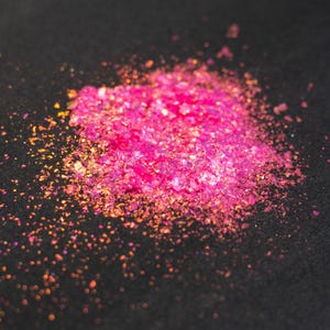 May include: A small pile of vibrant hot pink iridescent flakes with a reflective, metallic finish. The tiny, irregular fragments catch the light, displaying subtle shifts of orange and gold hues across the scattered, shimmering cluster of material.