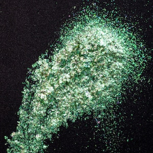 May include: A pile of loose, iridescent cosmetic flakes featuring a vibrant spectrum of emerald green, lime, and silver tones. The fine, irregular particles have a highly reflective, metallic finish that catches the light, creating a shimmering effect against the dark background.