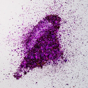May include: A pile of loose, iridescent purple flakes with a metallic finish. The flakes vary in size and shape, creating a textured, reflective cluster that radiates outward into smaller, scattered particles across a plain white surface.