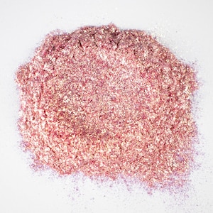 May include: A pile of loose, fine-grained cosmetic pigment powder in a vibrant metallic rose gold hue. The texture appears shimmering and reflective, with individual particles catching the light to create a multi-dimensional, sparkling effect.