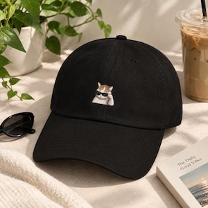 May include: A black baseball cap featuring a small, centered embroidery of a ginger and white cat wearing black sunglasses. The cap has a structured crown, a curved brim, and a button at the top, showcasing a clean, solid black fabric design.