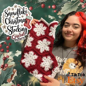 以下が含まれることがあります： 雪の結晶模様の赤い白のクロシェ編みクリスマスストッキング。ストッキングは人の手で持たれています。画像には「Snowflake Christmas Stocking Crochet Pattern」というテキストが含まれています。