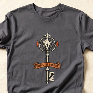 May include: A charcoal gray short-sleeve t-shirt featuring a graphic of an ornate key with a twenty-sided die head and a keyhole. An orange banner across the center of the key displays the text UNLOCK THE DUNGEON in black capital letters.