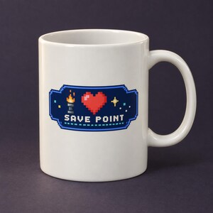 May include: A white ceramic mug featuring a pixel art graphic on the front. The design includes a blue rectangular border, a small torch, a red heart, a yellow star, and the text SAVE POINT in white pixelated letters centered on the mug.