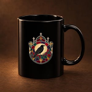 May include: A black ceramic mug featuring a central graphic of a black raven perched on a branch. The bird is framed by a circular, multi-colored stained glass pattern in shades of red, green, purple, and gold, set against a dark background.