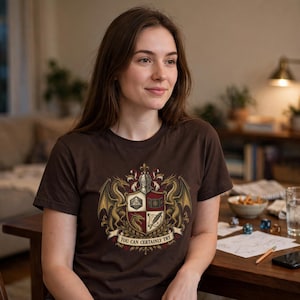 K&ouml;nnte beinhalten: Ein dunkelbraunes Kurzarm-T-Shirt mit einem detaillierten Wappen-Aufdruck. Das Motiv zeigt zwei goldene Drachen, die einen Schild mit einem zwanzigseitigen W&uuml;rfel, einem Buch, einem Drachenkopf und einer Feder flankieren. Der Text lautet "YOU CAN CERTAINLY TRY."