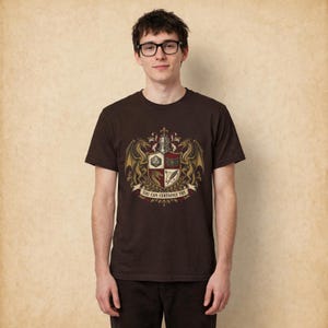K&ouml;nnte beinhalten: Ein dunkelbraunes Kurzarm-T-Shirt mit einem zentralen Wappenmotiv. Das Design zeigt zwei goldene Drachen, die einen Schild mit einem zwanzigseitigen W&uuml;rfel, einem Buch, einem Drachenkopf und einer Feder flankieren. Darunter steht der Text YOU CAN CERTAINLY TRY.