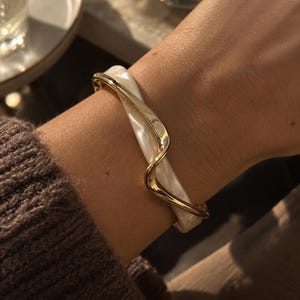 Może przedstawiać: Bransoletka typu bangle z gładką, białą, perłową bazą z żywicy, owiniętą polerowanym metalowym drutem w kolorze złota, który układa się w falisty wzór wokół obwodu opaski, tworząc warstwowy i przestrzenny wygląd biżuterii.
