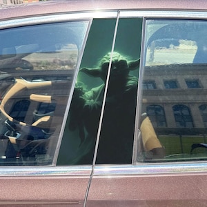 May include: A green, pointed-eared figure resembling a fictional character is positioned inside a vehicle, visible through the tinted glass of the rear passenger window. The figure has a wrinkled face and is wearing a light-colored robe.
