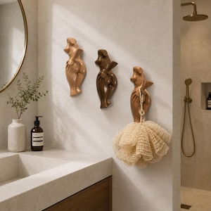 May include: Three wall-mounted hooks shaped like stylized human torsos in varying shades of tan and brown. The hooks are arranged vertically on a wall, with the rightmost hook supporting a beige mesh bath sponge hanging from its arm.