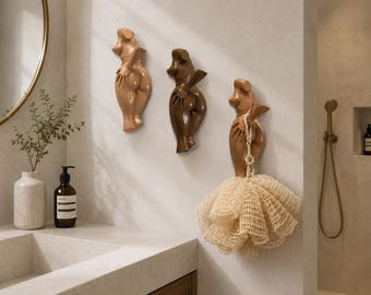 Könnte beinhalten: Drei Wandhaken in Form stilisierter menschlicher Torsi in verschiedenen Beige- und Brauntönen. Die Haken sind vertikal an einer Wand angebracht, wobei der rechte Haken einen beigen Netz-Badeschwamm trägt, der an seinem Arm hängt.