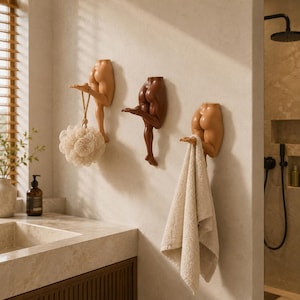 Op de afbeelding: Drie wandhaken in de vorm van menselijke onderlichamen in verschillende huidtinten. Elke haak heeft een gebeeldhouwd rompgedeelte en een enkel uitgestrekt been dat als hanger dient. Een beige badspons hangt aan de ene en een handdoek aan de andere.