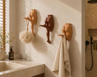 Könnte beinhalten: Drei Wandhaken in Form von menschlichen Unterkörpern in verschiedenen Hauttönen. Jeder Haken besteht aus einem geformten Rumpfbereich und einem einzelnen ausgestreckten Bein, das als Aufhängung dient. Ein beiger Badeschwamm und ein Handtuch hängen daran.