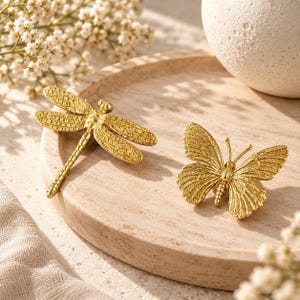 K&ouml;nnte beinhalten: Zwei goldfarbene Insektenfiguren aus Metall, eine in Form einer Libelle mit l&auml;nglichen Fl&uuml;geln und segmentiertem K&ouml;rper und die andere in Form eines Schmetterlings mit detaillierten Fl&uuml;gelmustern, auf einem runden Holztablett.