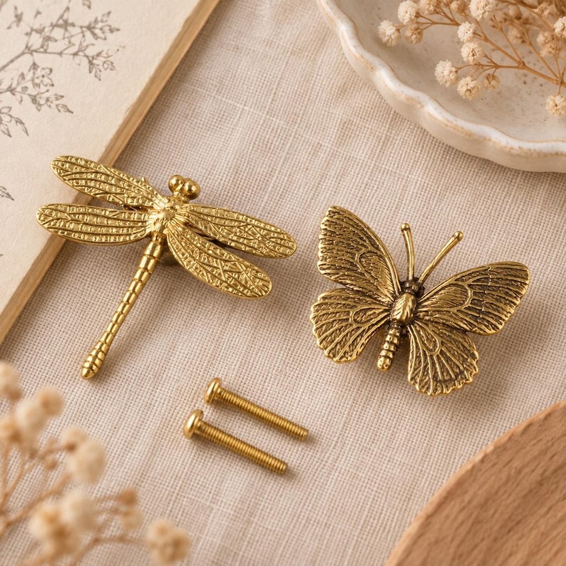 K&ouml;nnte beinhalten: Zwei messingfarbene M&ouml;belkn&ouml;pfe in Form einer Libelle und eines Schmetterlings, jeweils mit detaillierter Fl&uuml;gelstruktur. Zwischen den Kn&ouml;pfen befinden sich zwei kleine Metallschrauben zur Montage.
