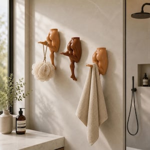 May include: Three wall-mounted hooks shaped like human lower torsos and legs in varying skin tones. Each hook features a bent leg extending outward to hold items, with a beige bath sponge hanging from the first and a beige towel from the third.