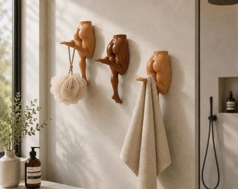Könnte beinhalten: Drei Wandhaken in Form von menschlichen Unterkörpern und Beinen in verschiedenen Hauttönen. Jeder Haken verfügt über ein abgewinkeltes Bein, das nach außen ragt, um Gegenstände zu halten, wobei ein beiger Badeschwamm und ein beiges Handtuch hängen.