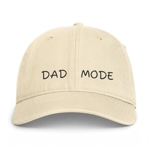 Op de afbeelding: Een beige honkbalpet met een gebogen klep en een gestructureerde kroon. Op de voorkant van de pet staat de tekst DAD MODE geborduurd in zwarte draad in een eenvoudig, afgerond lettertype. De pet heeft een klassieke constructie met zes panelen.