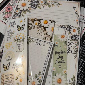 Könnte beinhalten: Ein Schreibwaren-Set mit Gänseblümchen- und Schmetterlingsillustrationen sowie schwarz-weißem Vichy-Muster. Es enthält einen linierten Notizblock, ein Lesezeichen mit dem Text A Brighter Day Awaits und einen Stickerbogen mit Daisy Vibes.