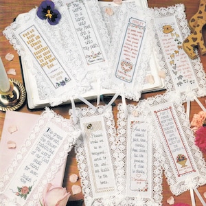 May include: A collection of eight rectangular fabric bookmarks featuring cross-stitched religious verses and illustrations, each bordered with white lace trim and finished with a white ribbon tie at the bottom, displayed on an open book.