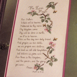 Peut inclure: Une broderie au point de croix sur tissu clair présentant le texte de la prière du Notre Père en lettres marron foncé. Le motif comprend des branches avec des feuilles vertes, des fleurs roses et deux petites abeilles, bordé d'un liseré violet.