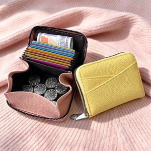 May include: A yellow zippered wallet with a textured exterior and an external pocket. Beside it, an open pink wallet reveals multiple card slots containing colorful cards, a rolled banknote, and several silver coins with crown motifs.