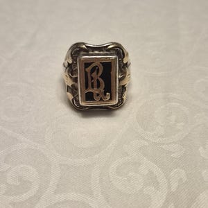 May include: A silver-toned ring featuring a rectangular face with a black inlay. The center of the inlay displays a raised, gold-colored monogram of the letters B and W in an ornate, gothic-style script. The band has decorative metal detailing.