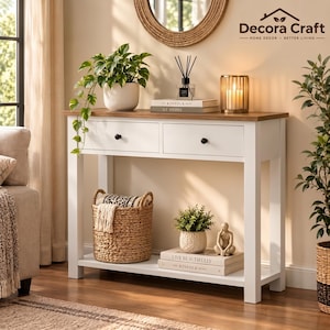May include: A white console table with a natural wood top, two drawers with black knobs, and a lower shelf. The table holds a potted plant, a reed diffuser, a candle, and books. A woven basket sits on the lower shelf with a small plant and figurine.