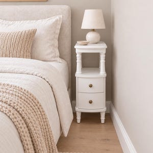 May include: A white bedside table with two drawers featuring round brass knobs, a lower shelf, and turned legs. A small white lamp with a beige shade and a small ceramic bowl sit on the top surface next to a bed with beige bedding and a knit throw.