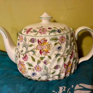 May include: A white ceramic teapot with a swirling, ribbed texture and a colorful floral pattern featuring pink, yellow, and purple blossoms with green leaves. The teapot includes a matching lid with a small knob and a curved handle and spout.