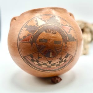 Puede incluir: Una vasija de barro de color terracota con un diseño circular pintado a mano en la parte frontal. La ilustración muestra un rostro estilizado con patrones geométricos en tonos negro, blanco y rojo apagado, enmarcado por un borde circular.