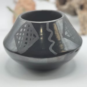May include: A small, polished black ceramic bowl with a wide, rounded base and a tapered opening. The exterior surface features matte grey hand-painted geometric patterns, including diamond shapes with grid lines and a vertical wavy line.