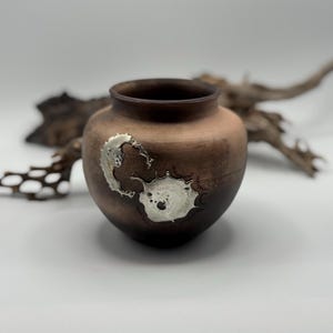 Może przedstawiać: Mały, okrągły, ciemnobrązowy wazon ceramiczny o matowym wykończeniu. Na zewnętrznej stronie znajdują się dwa wyraźne, nieregularne, metaliczne srebrne akcenty dekoracyjne o teksturze przypominającej stopiony metal.