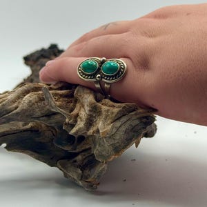 May include: A silver ring featuring two oval-shaped turquoise stones set horizontally. The metal setting has a twisted rope texture around the stones and a split shank band. The ring is displayed on a piece of weathered, textured driftwood.