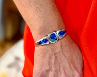 May include: A silver cuff bracelet featuring three inlaid sections of blue and green stone. The central inlay is rectangular, flanked by two smaller, elongated triangular inlays, all set within a polished silver metal band.