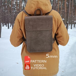 May include: A brown rectangular backpack with a top flap closure and shoulder straps. The image includes text that reads 5mm, pdf PATTERN, and YouTube VIDEO TUTORIAL. The backpack is worn over a mustard yellow hooded jacket and light-colored pants.