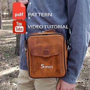 Peut inclure: Un sac bandoulière en cuir marron clair avec un rabat avant, un passepoil noir et une fermeture éclair. Le sac porte le texte 5mm, PDF PATTERN et YouTube VIDEO TUTORIAL. Il est porté sur une chemise grise et un pantalon beige en extérieur.
