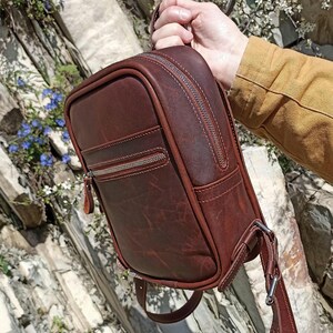 以下が含まれることがあります： 長方形のダークブラウンのレザー製バックパック。上部にハンドル、前面にファスナー付きポケットが付いています。バッグにはシルバーカラーの金属製ファスナーと、シルバーカラーのバックルが付いた調節可能なレザー製ショルダーストラップが備わっています。