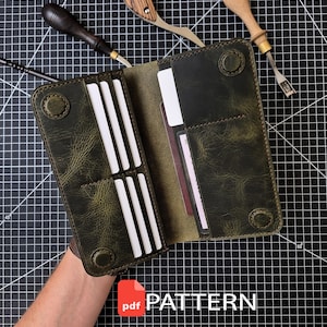 May include: An open olive green leather long wallet featuring multiple card slots, a large pocket for bills, and four circular snap closures. The wallet is displayed alongside three leatherworking tools on a black grid mat with a PDF PATTERN label.