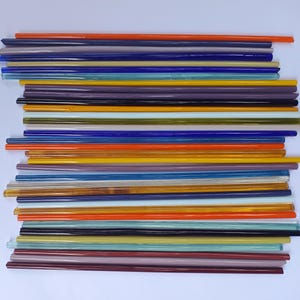 May include: A collection of long, thin, cylindrical glass rods arranged horizontally in a stack. The rods feature a variety of translucent and opaque colours, including shades of orange, blue, yellow, purple, brown, green, and clear glass.