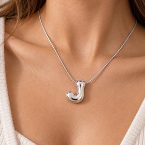 Puede incluir: Un collar de tono plateado con un colgante en forma de letra J tridimensional, lisa y abombada, suspendido de una cadena de serpiente fina y flexible. La superficie metálica de la letra tiene un acabado pulido que refleja la luz.